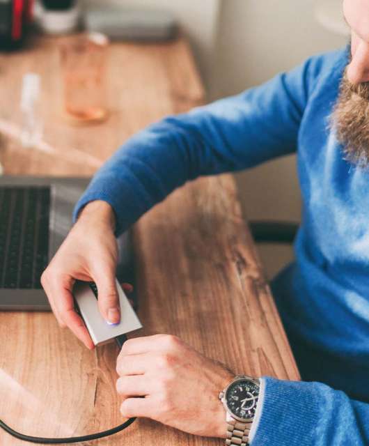 Man in blue sweater with brunette beard plugging external hard drive into laptop.