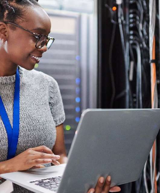 A woman holding a laptop smiles as she looks at a server