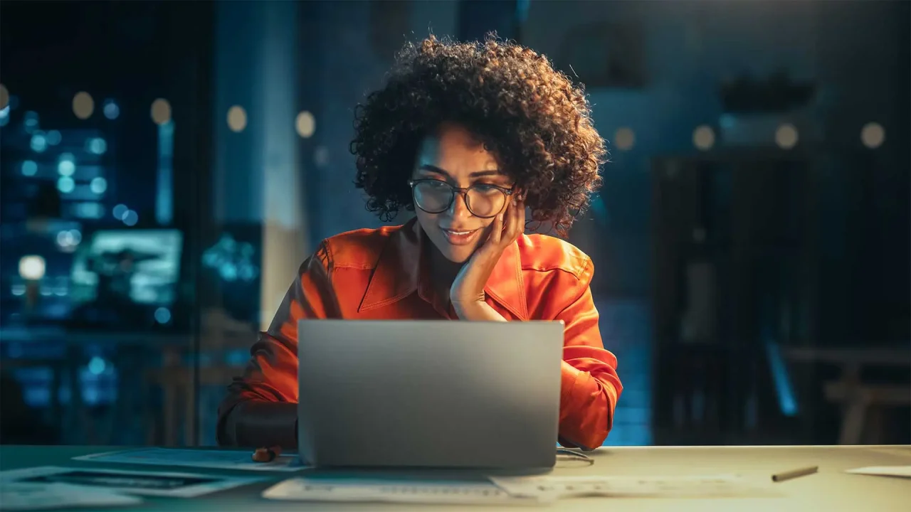 A woman sits at her laptop computer at night, looking intrigued